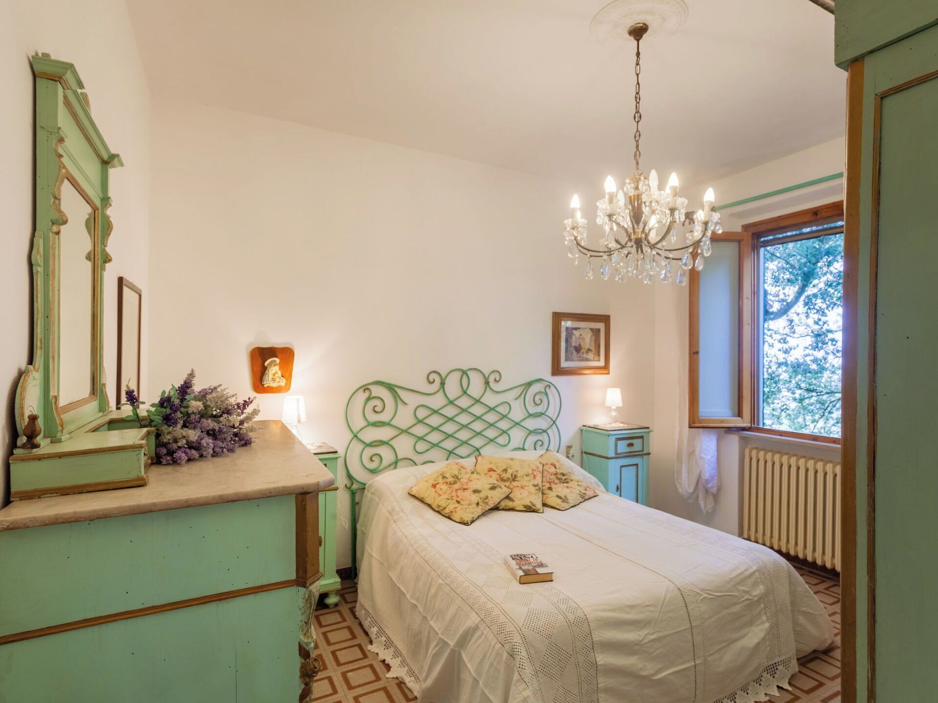 A bedroom with a bed, dresser, mirror, chandelier, and a window with a view.