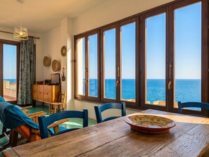 Ein Zimmer mit Holztisch, Stühlen, Meerblick durch große Fenster und einem Balkon mit Geländer.