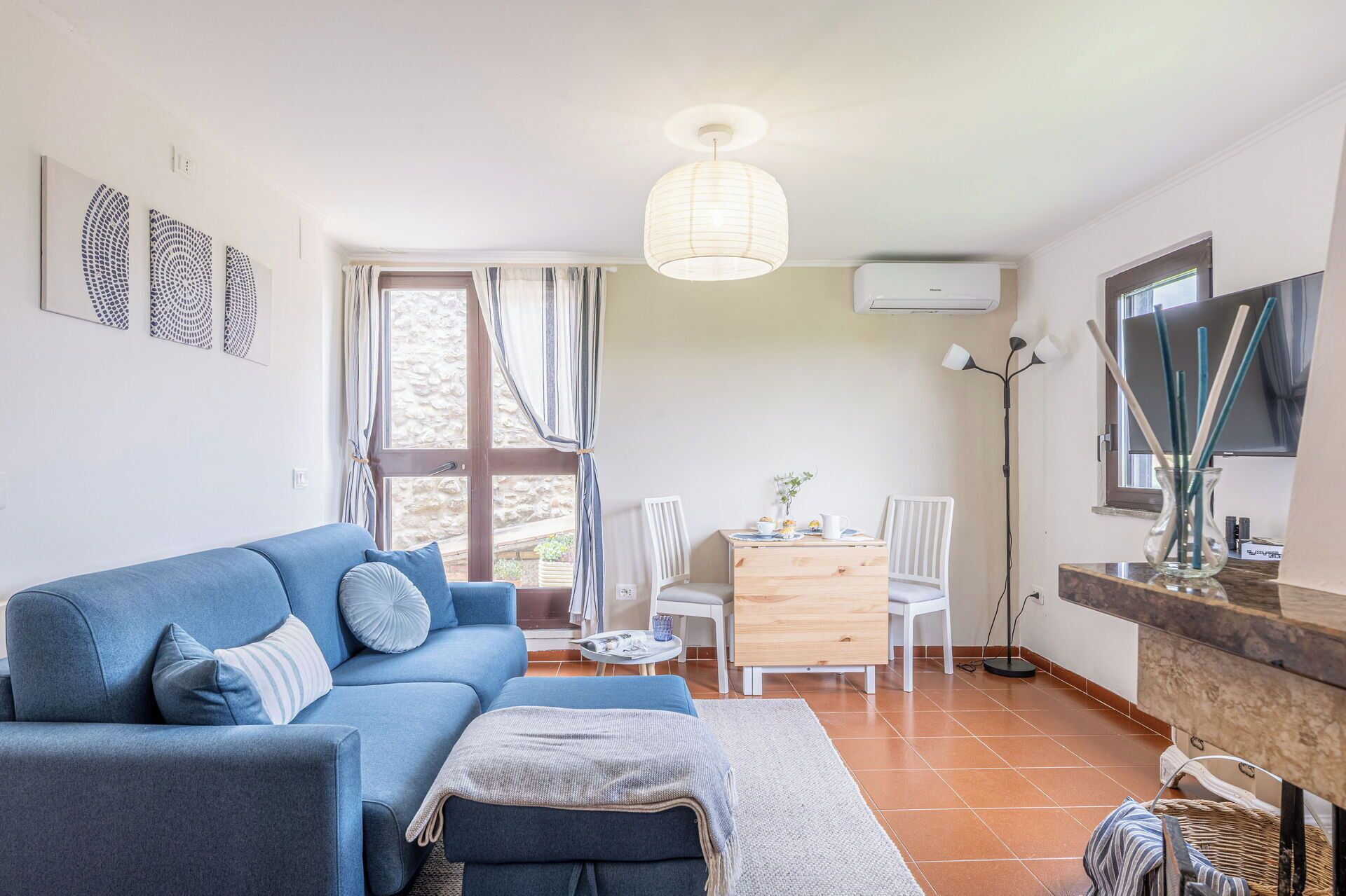 Un salon comprenant un canapé, une table à manger avec des chaises, un téléviseur et une grande fenêtre avec des rideaux.