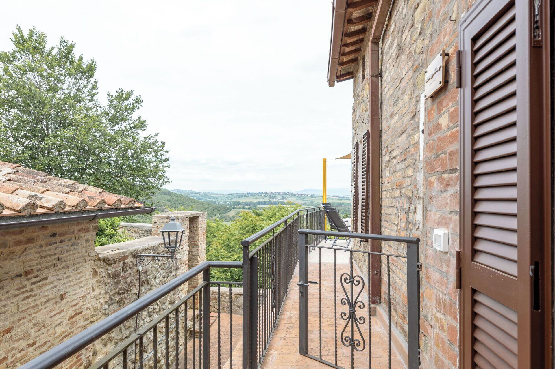 A balcony with a metal railing, a view of the countryside, and a brick building.