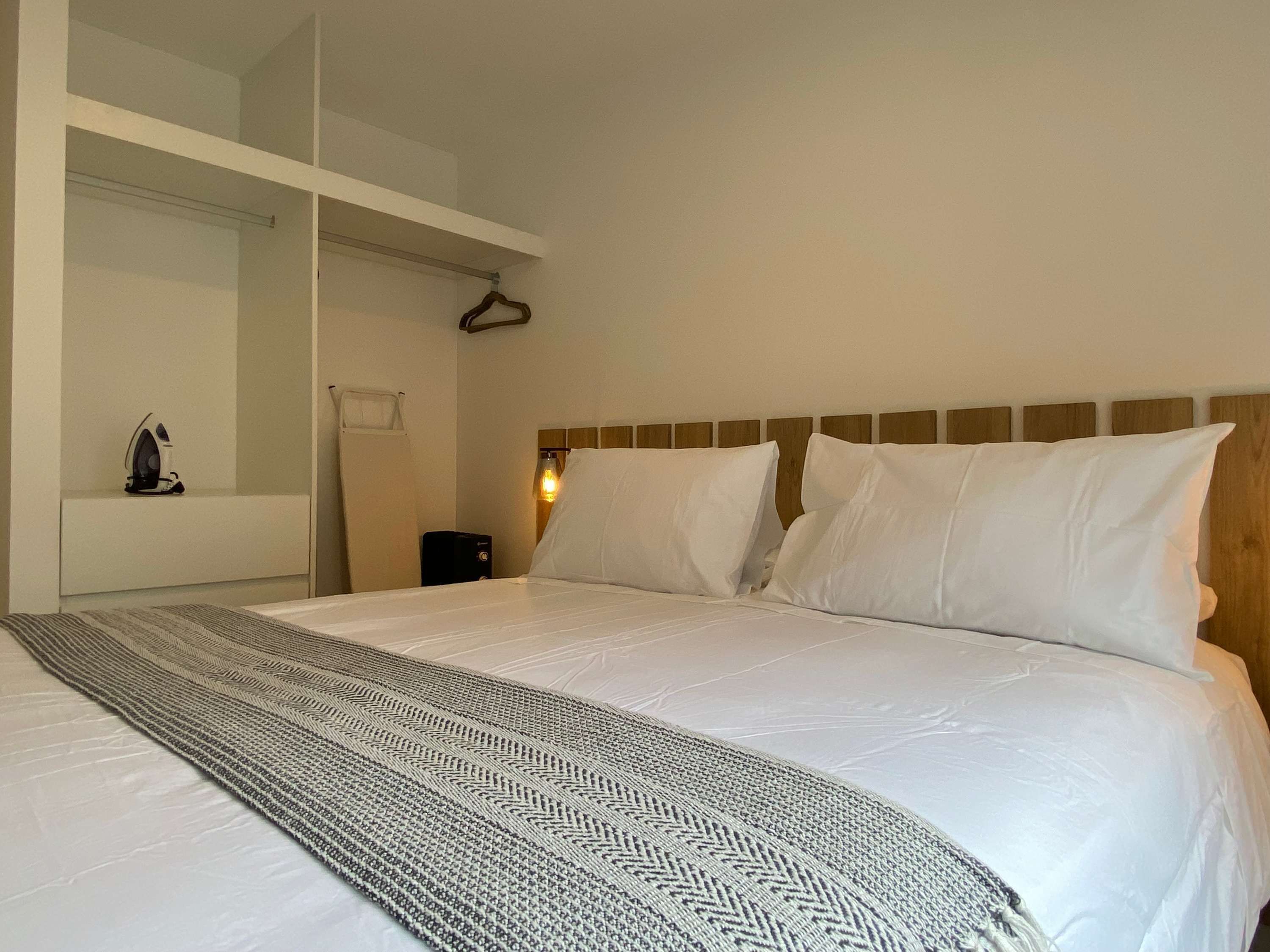 A hotel room with a bed, a bedside table, a hanging coat rack, and a wall-mounted heater.
