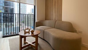 Una sala de estar moderna con un sofá, una mesita de madera y vistas a un paisaje urbano a través de amplias ventanas.