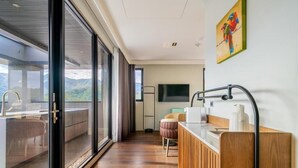 Una habitación de hotel moderna con un amplio ventanal corredero que da a un balcón, un tocador de madera con lavabo y un televisor de pantalla plana montado en la pared.