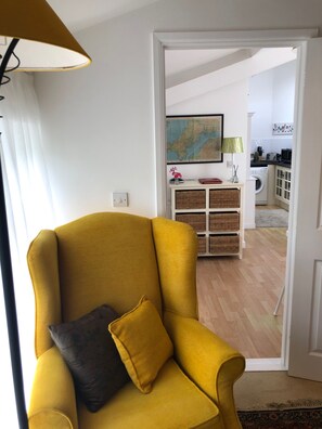 A yellow armchair with a dark cushion, a lamp, a map on the wall, a kitchen area with a washing machine, and a door leading to another room. - Coastal 1 Bed Flat - Sleeps 2 -patio - Parking (Boscastle)