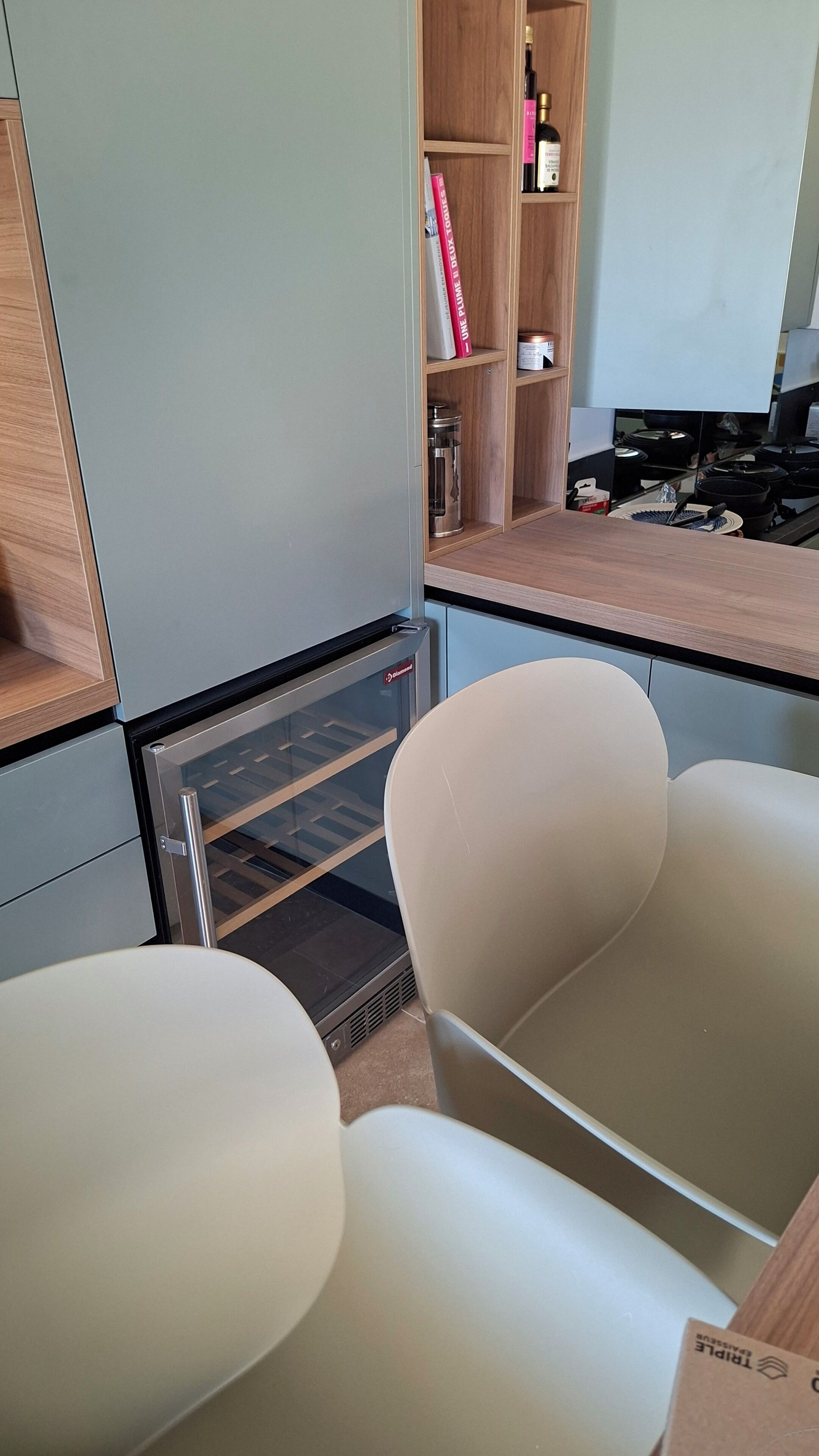 A kitchenette with a built-in oven, a coffee machine, and a small refrigerator. There are two white chairs and a wooden shelf with books and a bottle.