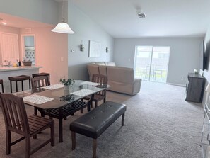 Ein Essbereich mit Glastisch, Holzsesseln und einer Bank. Eine Küchenzeile mit Arbeitsplatte und Barhocker ist ebenfalls vorhanden. Im Hintergrund ist eine Sitzecke mit Sofa und einem an der Wand befestigten Fernseher zu sehen.