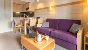 Een moderne hotelkamer met een paarse bank, een klein houten tafeltje met theepot en kopje, een laptop, en een kitchenette op de achtergrond.