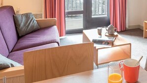 Een kamer met een paarse bank, een houten salontafel, een lamp, een raam met gordijnen en uitzicht op een balkon.