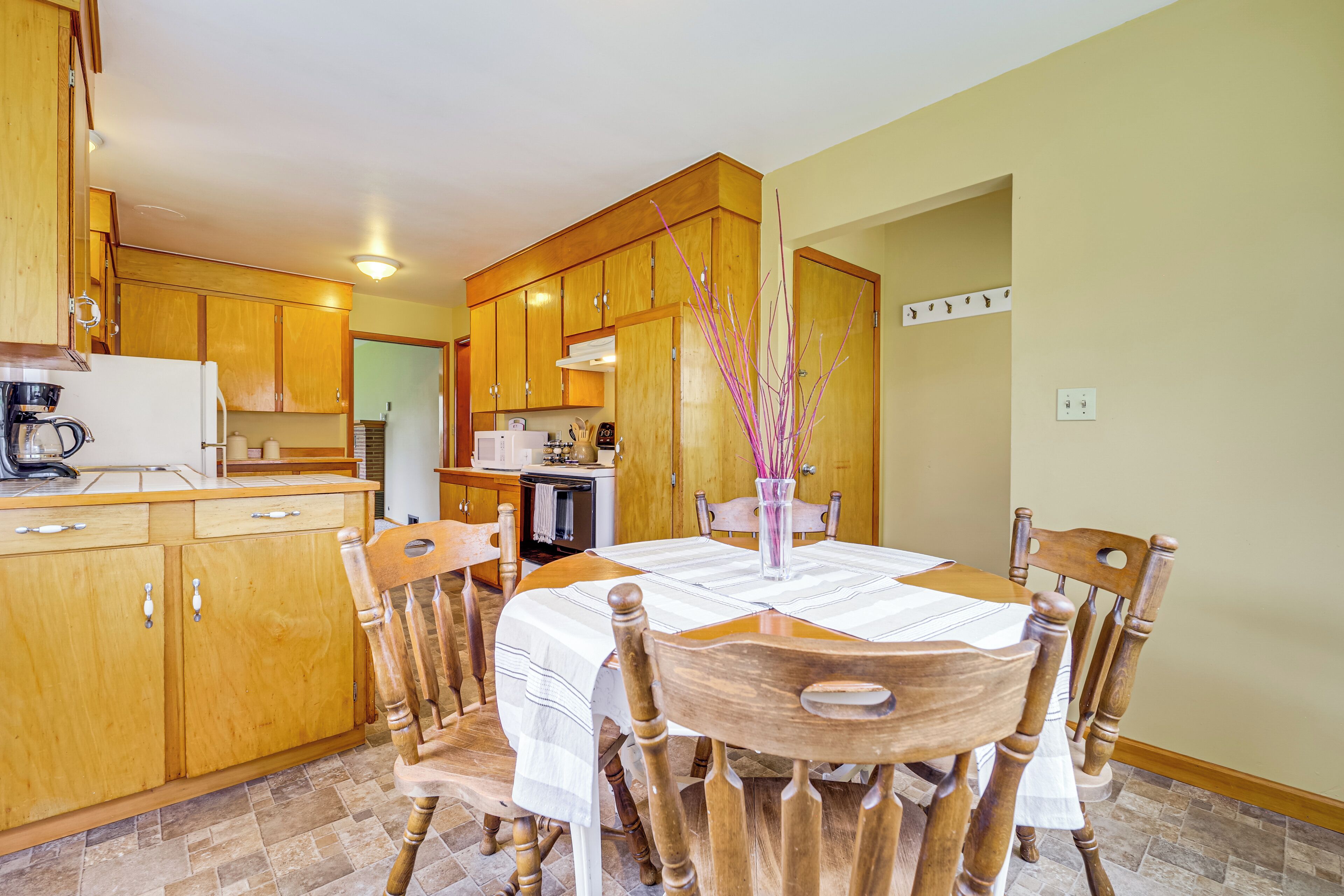 A dining area with wooden chairs and a table, a kitchen in the background, and a vase with a plant on the table.