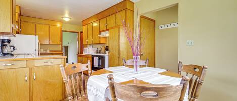 A dining area with wooden chairs and a table, a kitchen in the background, and a vase with a plant on the table.