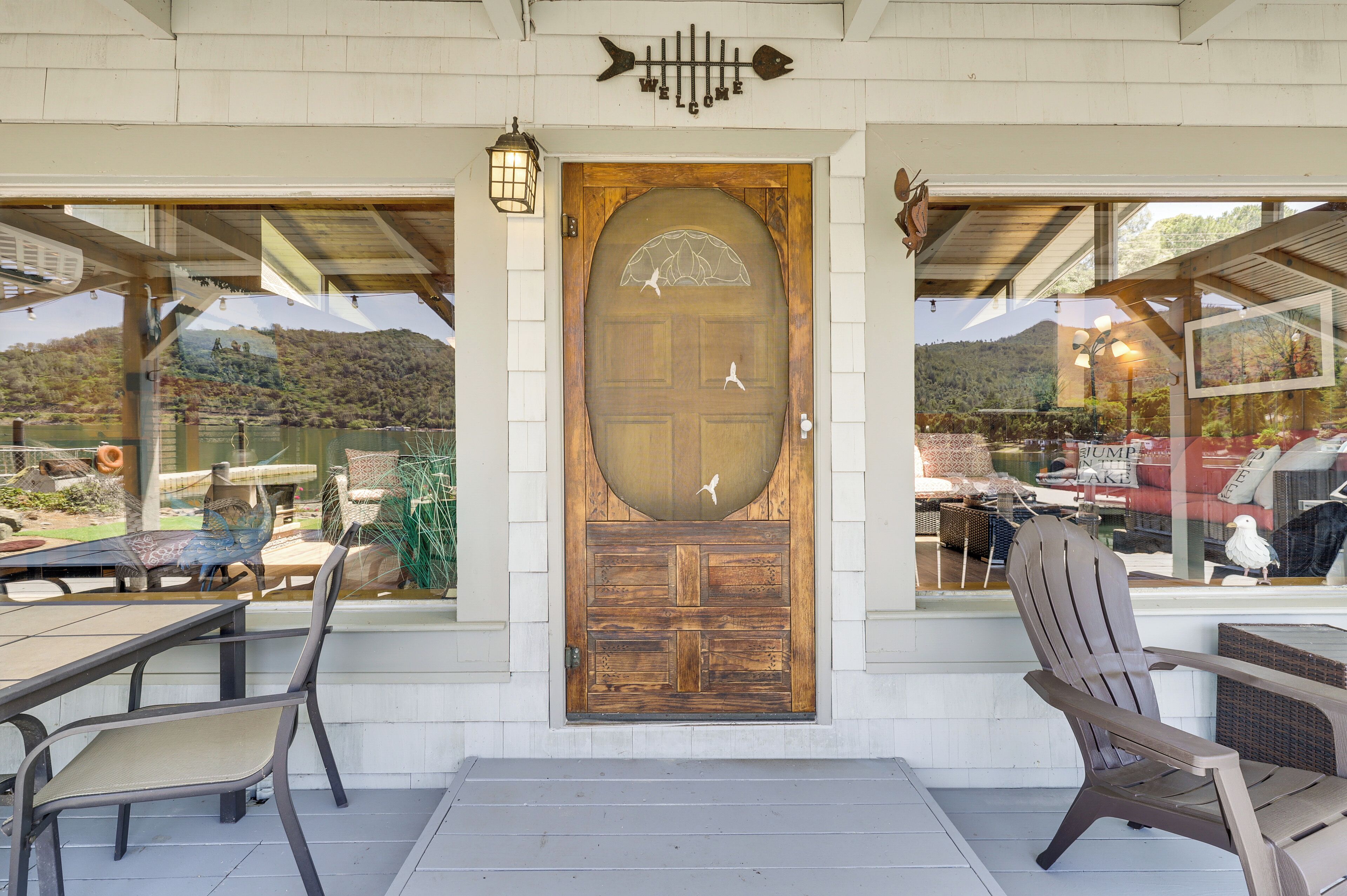 A wooden door with a welcome sign above, a view of a mountain through the window, and outdoor seating with chairs.