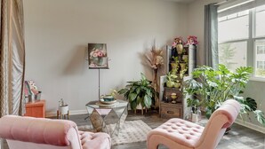 Un salon avec une grande fenêtre, une table basse ronde, deux fauteuils roses et diverses plantes.