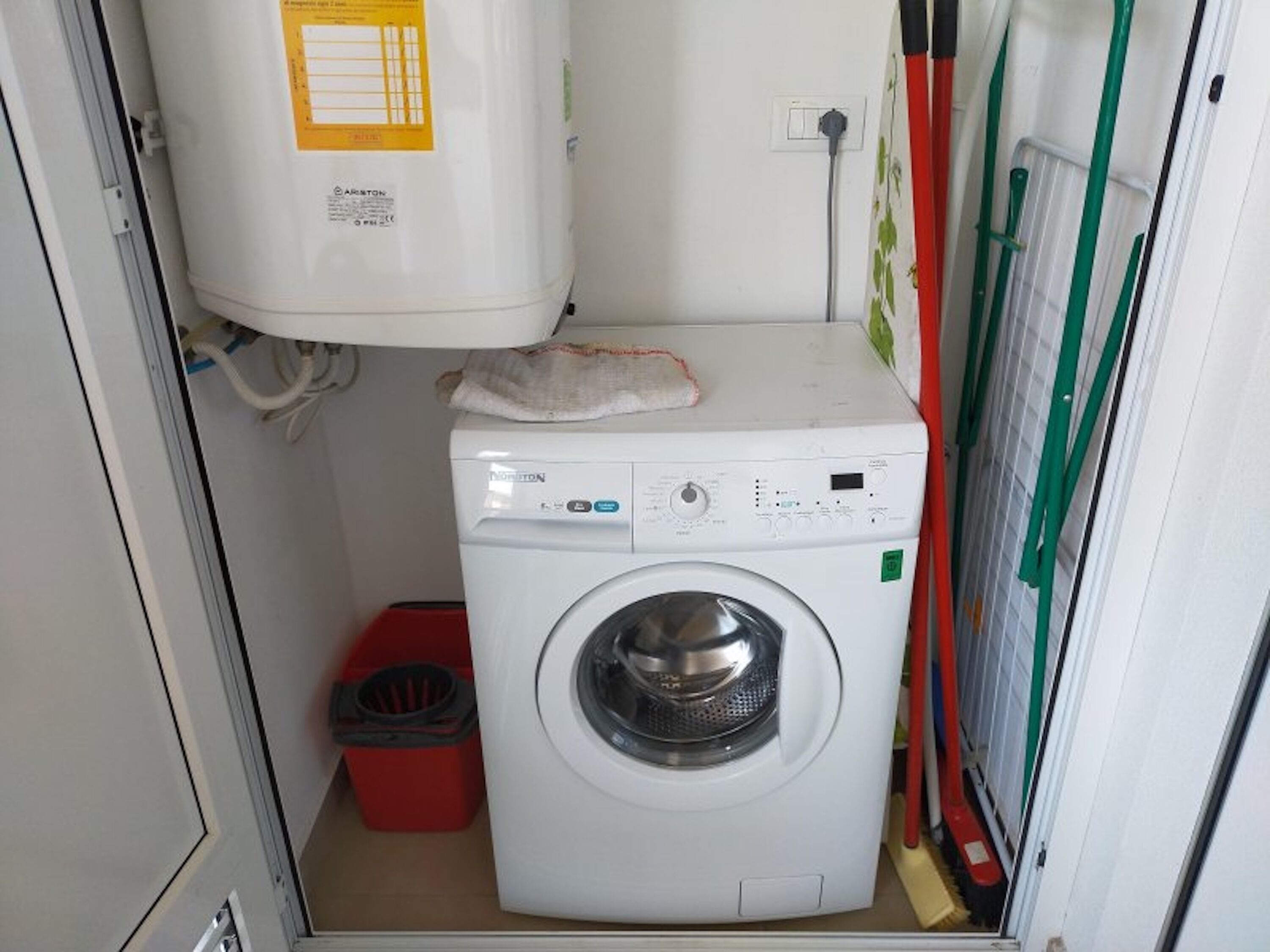 Une petite buanderie équipée d’un lave-linge à chargement frontal, d’un chauffe-eau et d’une poubelle rouge.