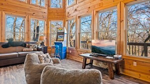 A cabin interior with a large window, a sofa, a chair, a table with a painting, and a gaming machine. - Sleeps 14! Hot Tub*Resort Amenities*Arcade*Games (Gatlinburg)