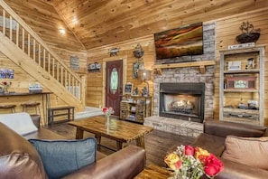 L’intérieur d’une cabane en rondins, avec une cheminée, un escalier en bois et un coin salon confortable.
