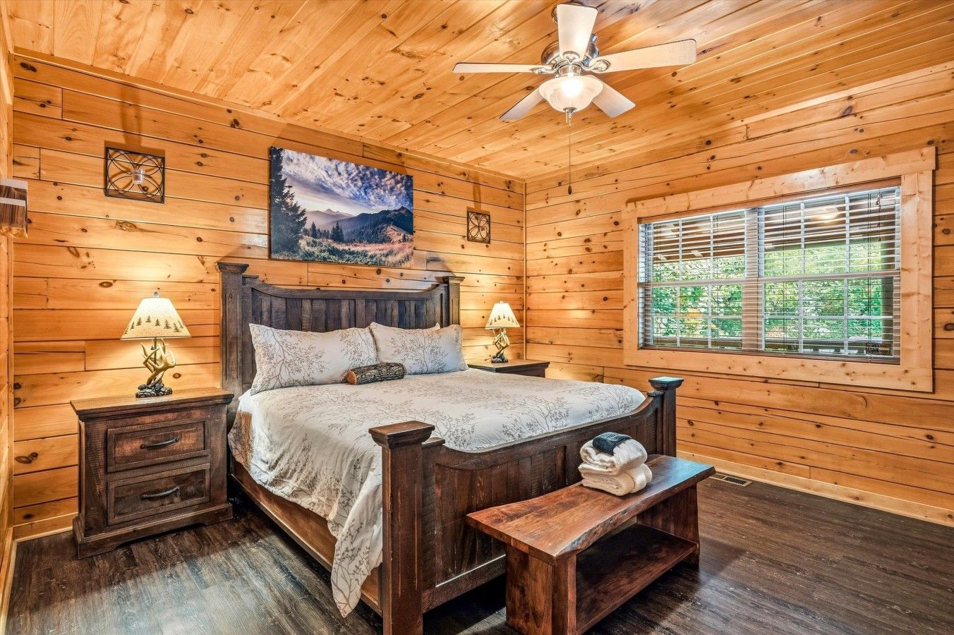 A wooden cabin bedroom with a bed, nightstands, a bench, a ceiling fan, a painting, and a window with blinds.