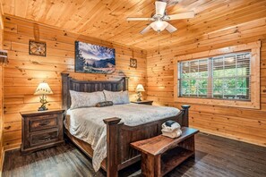 A wooden cabin bedroom with a bed, nightstands, a bench, a ceiling fan, a painting, and a window with blinds. - 4BR w/MTN Views! Hot Tub*Game Room*Resort*Quiet! (Gatlinburg)