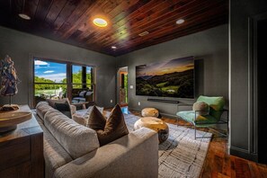 Ein modernes Wohnzimmer mit einem großen Flachbildfernseher, einem Sofa und Blick auf eine Landschaft durch ein großes Fenster.
