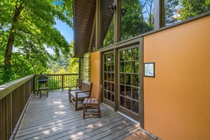 Eine Holzterrasse mit zwei Stühlen und einem Grill, mit Blick auf ein bewaldetes Gebiet.