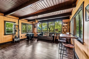 Ein geräumiger Raum mit Holzbalkendecke, großen Fenstern mit Jalousien, einer Couch, einem Esstisch mit Hocker und einer an der Wand stehenden Leiter.