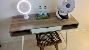 Ett skrivbord i trä med en vit låda, en liten pall, en skrivbordslampa, två vattenflaskor och en fläkt.