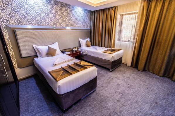 A hotel room with two beds, a headboard, a nightstand, a bedside table, and a window with curtains. - Al Jaber Towers Hotel - Makkah (Makkah)