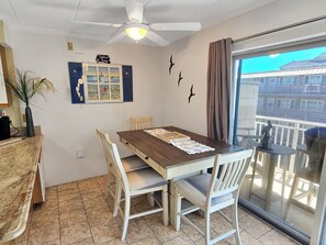 En matplats med ett träbord, vita stolar, en takfläkt och utsikt över byggnader genom en glasdörr.