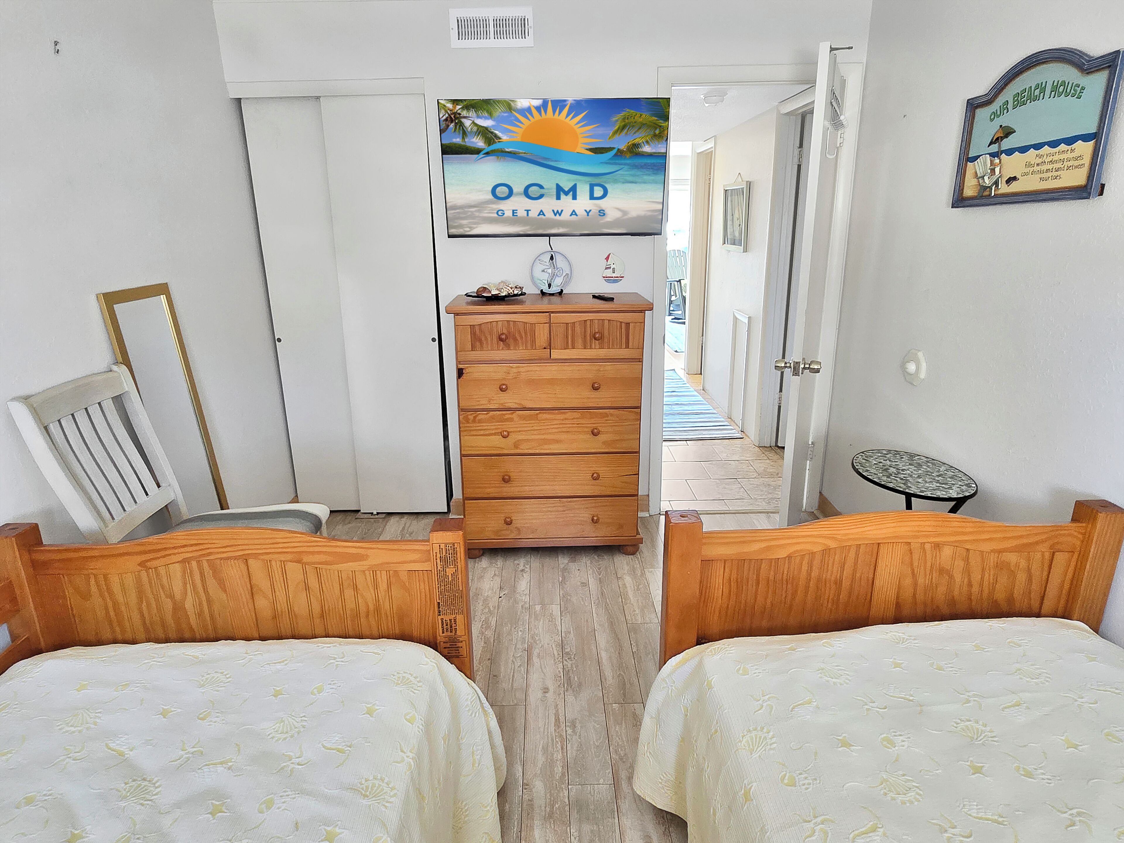 A bedroom with two wooden beds, a dresser, a painting of a beach, and a sign that reads 'Our Beach House.'