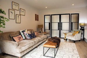 Un soggiorno con un divano, una poltrona, un poggiapiedi e cuscini decorativi. Alle pareti sono presenti opere d'arte incorniciate e una porta finestra decorativa.