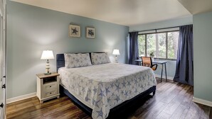 A bedroom with a bed, two bedside tables with lamps, a desk, and a window with curtains. - Sunset Rentals | Treetops 6101 | Dog Friendly (Hilton Head Island)
