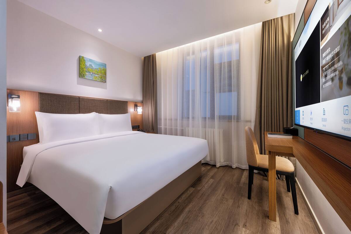 A hotel room with a bed, a desk, a chair, a TV, and a painting on the wall.