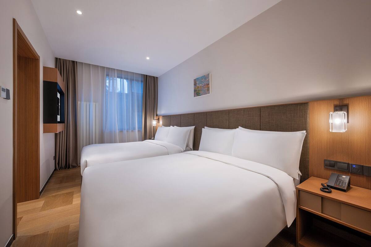 A hotel room with two beds, a wooden headboard, a bedside table with a phone, and a wall-mounted light.