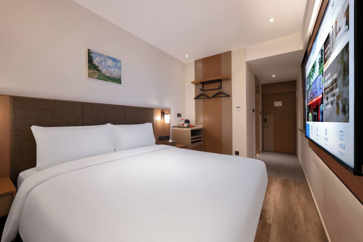 A hotel room with a large bed, a desk with a chair, a TV mounted on the wall, and a door leading to another room.