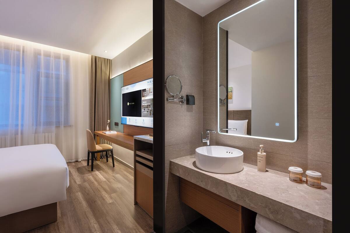 A modern hotel room with a bed, a desk, a large mirror, a round mirror, a sink, and a vanity.