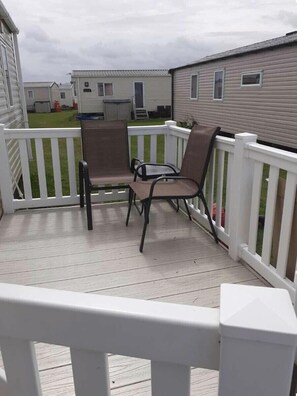 Een overdekt terras buiten, met een witte balustrade, twee stoelen en zicht op stacaravans in de verte.
