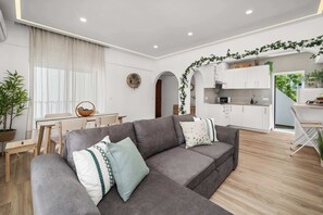 Uma sala de estar moderna com um sofá modular cinza, uma área de jantar com mesa e cadeiras de madeira, e uma cozinha com armários brancos e uma planta pendente.
