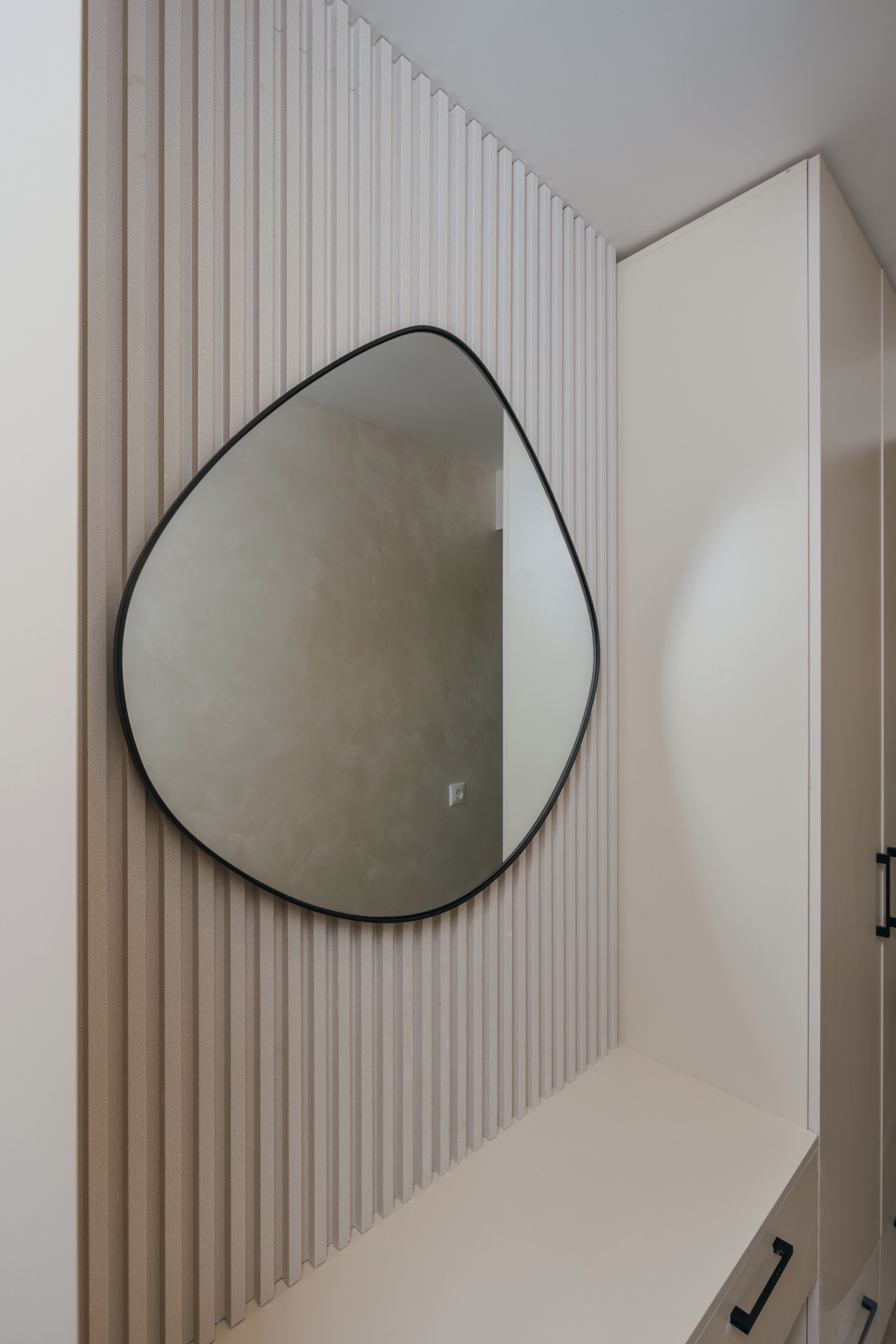 A room with a wall-mounted mirror, a white cabinet, and a vertical panel design.