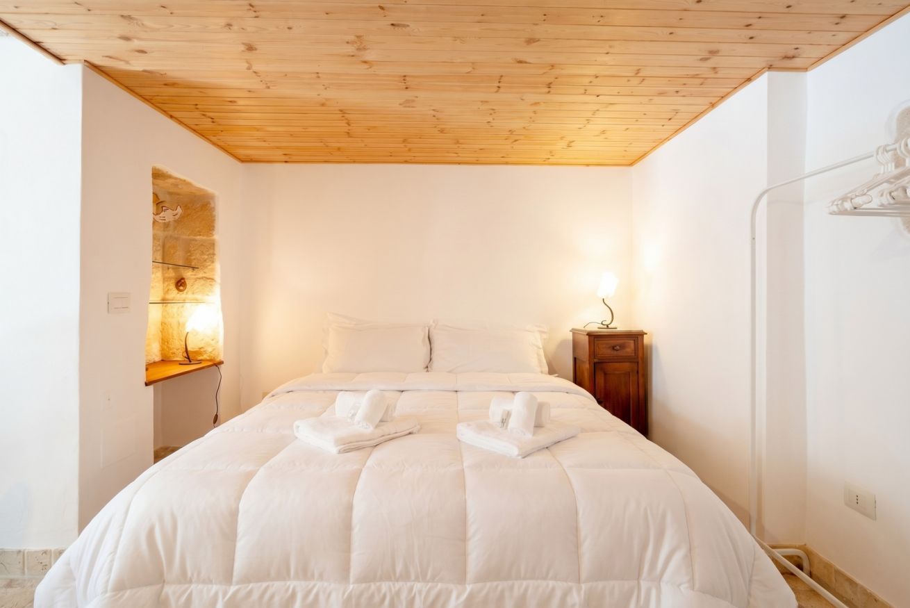 Une chambre avec un plafond en bois, un lit avec une literie blanche, une table de chevet et un luminaire fixé au mur.