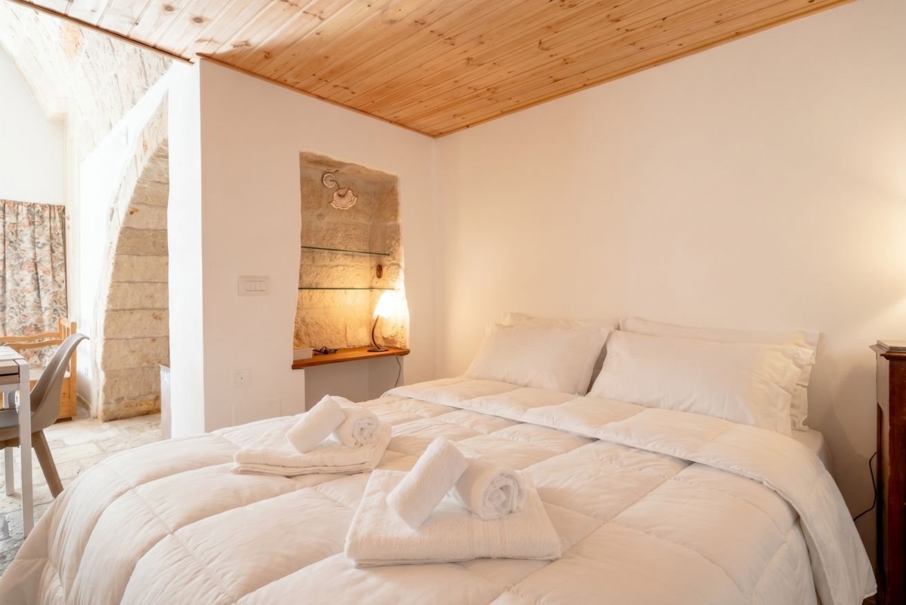 A bedroom with a wooden ceiling, a bed with white bedding, and a stone wall with a light fixture.
