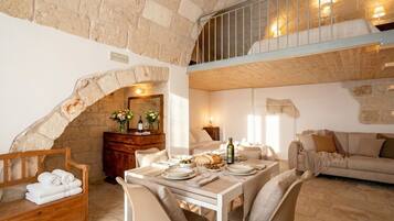 A rustic room with a stone wall, a wooden bench with towels, a dining table set for two, and a sofa.