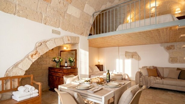 Une pièce rustique avec un mur en pierre, un banc en bois sur lequel sont posées des serviettes, une table à manger dressée pour deux personnes et un canapé.