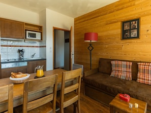Ein gemütliches Zimmer mit einer Holzwand, einer Couch mit Karokissen, einem Esstisch mit Stühlen, einer Küchenzeile inklusive Mikrowelle und einer roten Stehlampe.