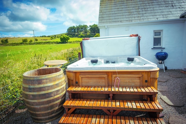 A hot tub with a wooden bench, a wooden barrel, and a barbecue grill outside a house with a white exterior.