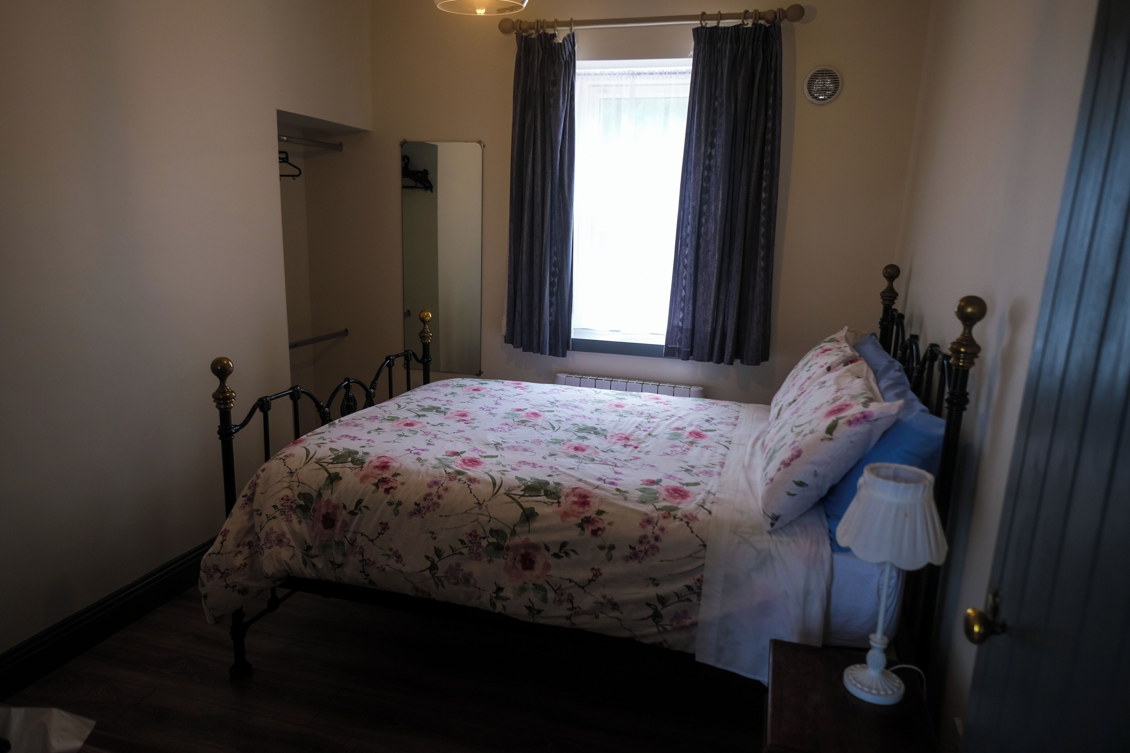 A bedroom with a bed, a bedside table with a lamp, a window with curtains, a door, and a mirror on the wall.