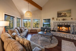 Ein Wohnzimmer mit Kamin aus Stein, einer Sofagarnitur, einem Couchtisch und großen Fenstern mit Jalousien.