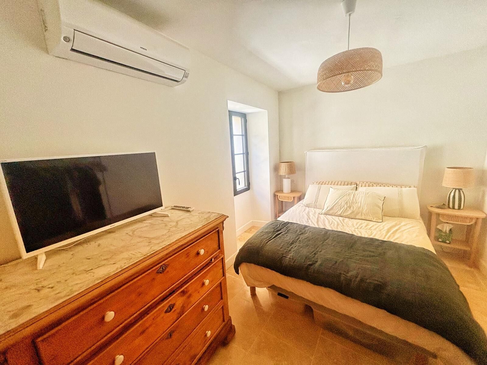 Een slaapkamer met een bed, een kapstok, een televisietoestel, een raam en een hangende lamp.