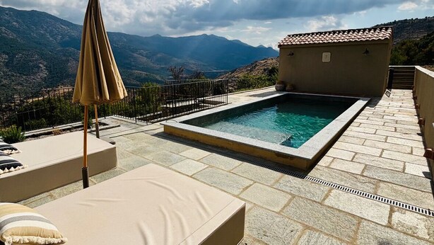A pool area with a lounge chair and umbrella, surrounded by stone tiles and a view of mountains. - Stone house with swimming pool in the heart of the Corsican mountains. Near Corte (Corscia)