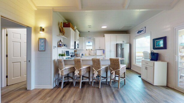A kitchen with white cabinets, a central island with wicker chairs, a refrigerator, and a view through a window. - Bolivar Bungalow: 1  BR, 1.5  BA House in Crystal Beach, Sleeps 5 (Crystal Beach)