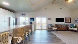 A spacious living area with a large sliding glass door to a deck, a kitchen with white cabinets, wicker furniture, and a flat-screen TV mounted on the wall. - Bolivar Bungalow: 1  BR, 1.5  BA House in Crystal Beach, Sleeps 5 (Crystal Beach)