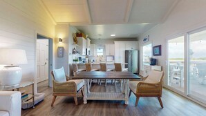 A spacious dining area with a large wooden table and wicker chairs, a kitchen with white cabinets, and a view of the beach through large windows. - Bolivar Bungalow: 1  BR, 1.5  BA House in Crystal Beach, Sleeps 5 (Crystal Beach)